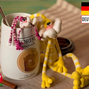 May include: Two crocheted animal toys, one pink and one yellow, with white accents, are sitting on a white mug with the words "Forest Animals" and "The owls" printed on it. The mug is on a brown and white striped surface.