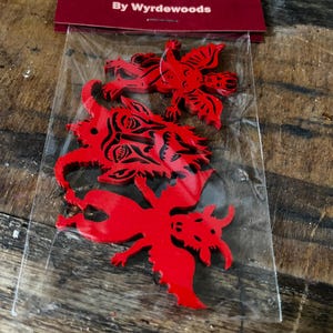 K&ouml;nnte beinhalten: Drei rote Holzausschnitte mit teufelsartigen Figuren und schwarzen Tintendetails, verpackt in einer durchsichtigen Plastikt&uuml;te mit einer kastanienbraunen Kopfkarte mit der Aufschrift Wee Devils By Wyrdewoods. Enth&auml;lt ein geh&ouml;rntes Gesicht und zwei gefl&uuml;gelte Wesen.