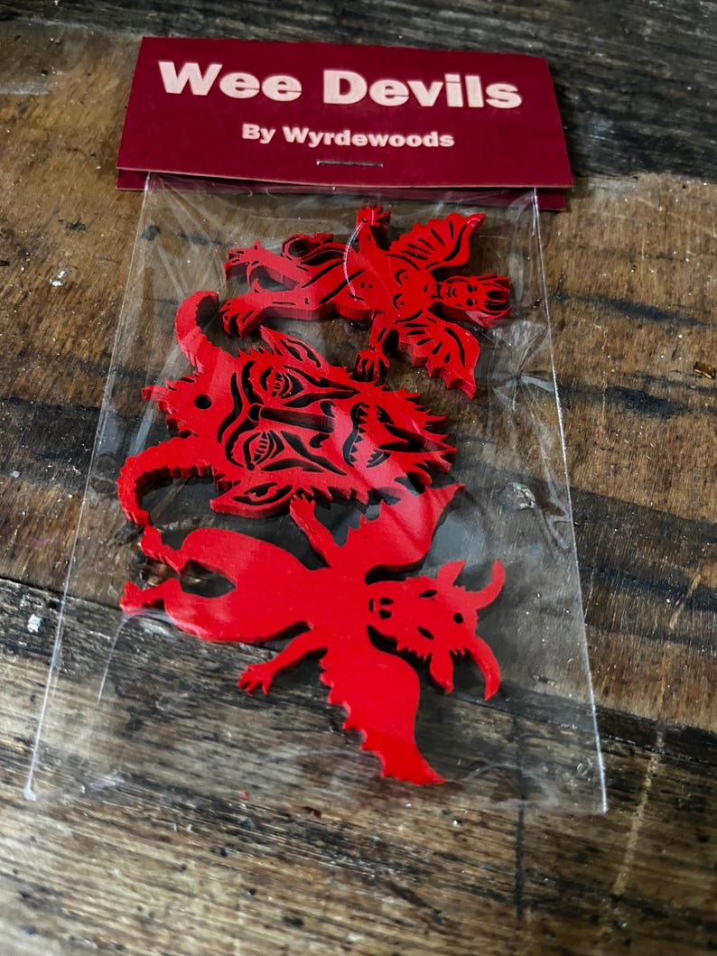 K&ouml;nnte beinhalten: Drei rote Holzausschnitte mit teufelsartigen Figuren und schwarzen Tintendetails, verpackt in einer durchsichtigen Plastikt&uuml;te mit einer kastanienbraunen Kopfkarte mit der Aufschrift Wee Devils By Wyrdewoods. Enth&auml;lt ein geh&ouml;rntes Gesicht und zwei gefl&uuml;gelte Wesen.