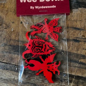 K&ouml;nnte beinhalten: Ein durchsichtiger Plastikbeutel mit drei flachen, roten, teufelsf&ouml;rmigen Ausschnitten. Der Beutel ist oben mit einem dunkelroten Pappschild verschlossen, das den wei&szlig;en Text "Wee Devils" und "By Wyrdewoods" tr&auml;gt. Die Ausschnitte zeigen detaillierte schwarze Linien.
