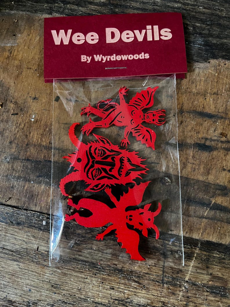 K&ouml;nnte beinhalten: Ein durchsichtiger Plastikbeutel mit drei flachen, roten, teufelsf&ouml;rmigen Ausschnitten. Der Beutel ist oben mit einem dunkelroten Pappschild verschlossen, das den wei&szlig;en Text "Wee Devils" und "By Wyrdewoods" tr&auml;gt. Die Ausschnitte zeigen detaillierte schwarze Linien.