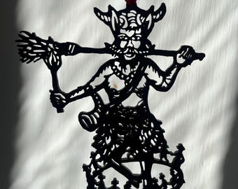 Könnte beinhalten: Ein schwarzer, flacher, lasergeschnittener Anhänger, der eine gehörnte Figur mit Schnurrbart zeigt, die einen Besenstiel über den Schultern trägt. Die Figur steht auf einem runden Sockel mit tanzenden Silhouetten und hängt an einem roten Band.