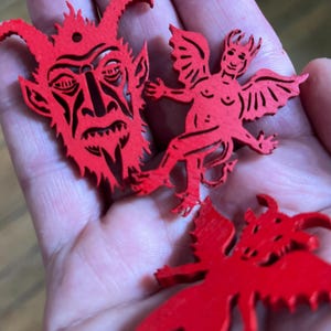 K&ouml;nnte beinhalten: Drei leuchtend rote, flache, lasergeschnittene Holzornamente. Ein Teil ist ein detailliertes Teufelsgesicht mit H&ouml;rnern und einem kleinen Loch an der Oberseite. Die anderen beiden Teile sind gefl&uuml;gelte Teufelsfiguren mit spitzen Schw&auml;nzen.