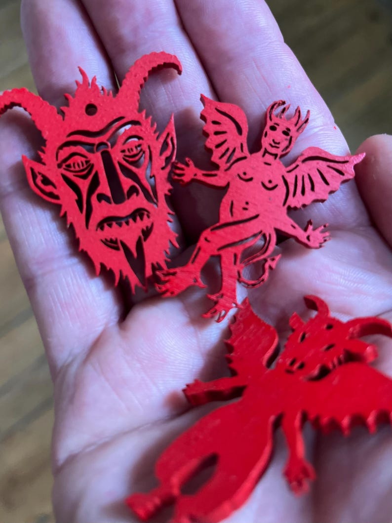 K&ouml;nnte beinhalten: Drei leuchtend rote, flache, lasergeschnittene Holzornamente. Ein Teil ist ein detailliertes Teufelsgesicht mit H&ouml;rnern und einem kleinen Loch an der Oberseite. Die anderen beiden Teile sind gefl&uuml;gelte Teufelsfiguren mit spitzen Schw&auml;nzen.