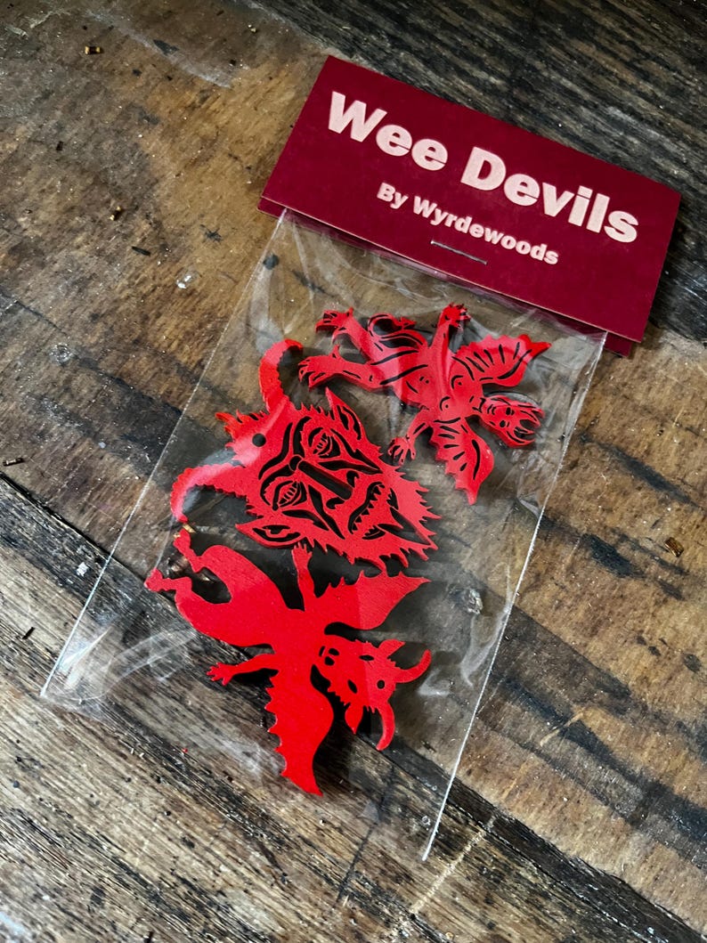 K&ouml;nnte beinhalten: Ein durchsichtiger Plastikbeutel mit drei roten Holzausschnitten von Teufelsfiguren mit schwarzen Druckdetails. Die Verpackung hat eine dunkelrote Kopfkarte mit dem wei&szlig;en Text Wee Devils By Wyrdewoods auf der Vorderseite.