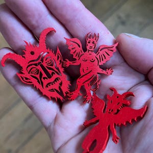 K&ouml;nnte beinhalten: Drei leuchtend rote Holzanh&auml;nger mit filigranen Ausschnitten. Ein Anh&auml;nger zeigt ein geh&ouml;rntes Teufelsgesicht, ein weiterer eine gefl&uuml;gelte Figur mit H&ouml;rnern und der dritte ein gefl&uuml;geltes Wesen mit spitzem Kopf und Schwanz, alle mit Gravuren.