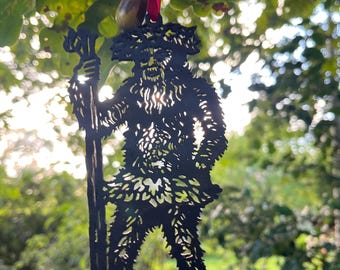 Könnte beinhalten: Schwarze Metallaussparung einer Waldkreatur mit Stab, aufgehängt an einem roten Band mit Eicheln. Die detaillierte Silhouette zeigt einen Bart und Kopfbedeckung aus Blättern, vor einem Hintergrund aus grünem Laub.