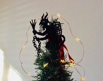Könnte beinhalten: Eine schwarze Silhouette eines Teufels, der eine Kette und einen Korb hält, als Christbaumspitze verwendet. Der Baum ist mit einer Lichterkette aus Kupferdraht mit sternförmigen Glühbirnen geschmückt.
