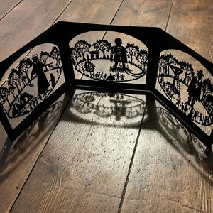 May include: A black paper cut-out diorama depicting a scene in a park. The diorama is made up of three panels that fold out to reveal the scene. The scene includes trees, people, and a path.