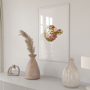 May include: A white framed print with a gold circle featuring pink and white flowers. The print is hanging on a white wall above a wooden dresser with a vase of dried flowers, a decorative object, and a lamp.