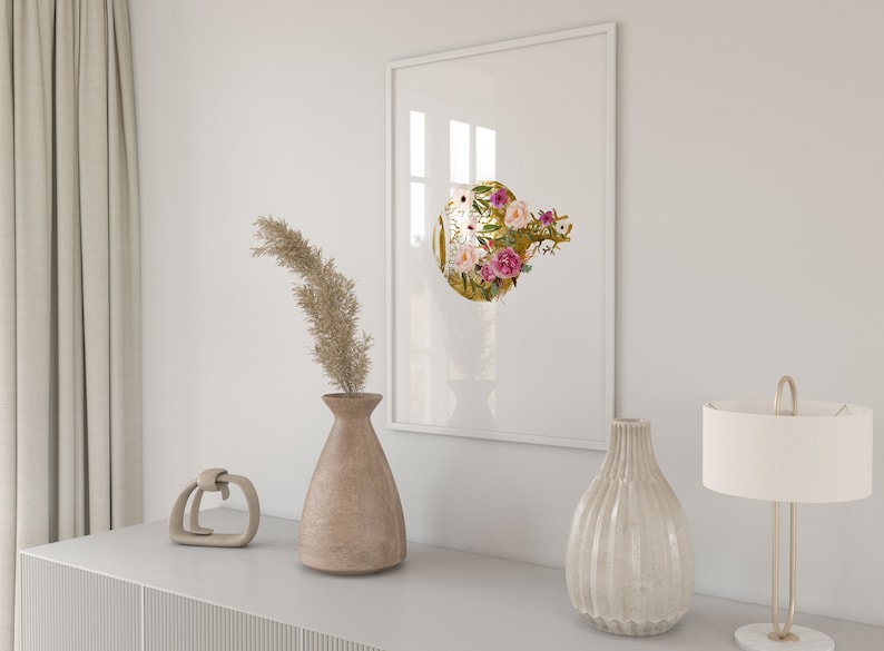 May include: A white framed print with a gold circle featuring pink and white flowers. The print is hanging on a white wall above a wooden dresser with a vase of dried flowers, a decorative object, and a lamp.