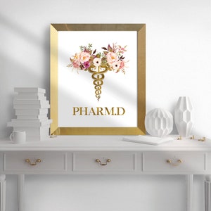 May include: A gold framed print with a white background. The print features a gold caduceus symbol with pink and white flowers. The text "PHARM.D." is written below the symbol.