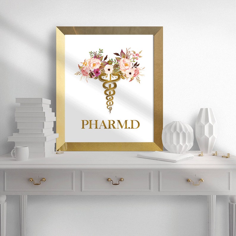 May include: A gold framed print with a white background. The print features a gold caduceus symbol with pink and white flowers. The text "PHARM.D." is written below the symbol.