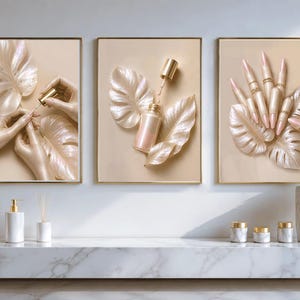 Könnte beinhalten: Drei gerahmte Wandbilder in beigen Farbtönen mit irisierenden Monstera-Blättern, Kosmetikflaschen und manikürten Fingern. Die Kunstwerke hängen über einem weißen Marmorregal mit passenden Badezimmeraccessoires und verschiedenen Behältern.