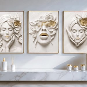 May include: A set of three rectangular wall art prints featuring white relief-style portraits of faces adorned with gold-accented flowers and lips. The artwork is displayed above a white marble counter with various white and gold bathroom accessories.