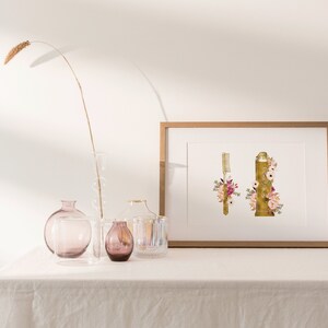 May include: A framed print of a gold toothbrush and toothpaste tube decorated with pink and white flowers. The print is on a white table with a vase of dried grass and other decorative vases.