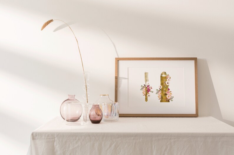 May include: A framed print of a gold toothbrush and toothpaste tube decorated with pink and white flowers. The print is on a white table with a vase of dried grass and other decorative vases.