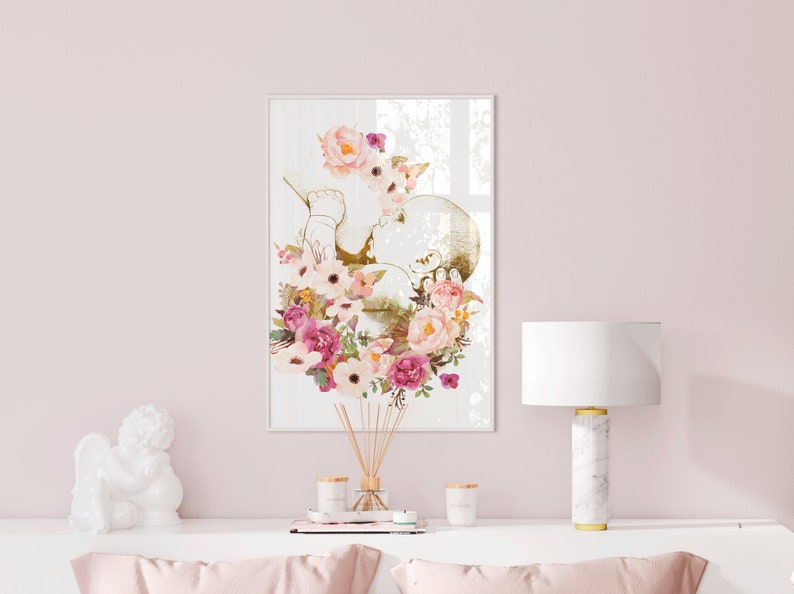 May include: A white framed print with a gold outline of a baby in a floral wreath. The flowers are pink, white, and cream.
