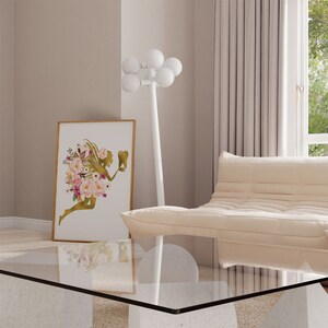 May include: A gold silhouette of a fairy with flowers and a tooth in her hand, framed in a gold frame, hanging on a light beige wall. A white floor lamp with multiple round white shades is in the background.