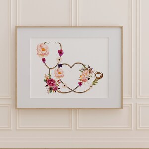May include: A gold stethoscope with a heart shape formed by the tubing, decorated with pink, white, and purple flowers. The stethoscope is framed in a white frame with a wood border.