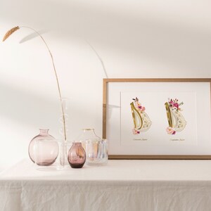 May include: A framed print with two gold letters, "D" and "D", decorated with pink and white flowers. The letters are on a white background with the names "Donovan Joseph" and "Sophia Rose" written below each letter. The print is sitting on a white table with a vase and other decorative items.