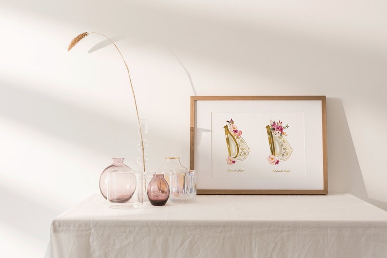May include: A framed print with two gold letters, "D" and "D", decorated with pink and white flowers. The letters are on a white background with the names "Donovan Joseph" and "Sophia Rose" written below each letter. The print is sitting on a white table with a vase and other decorative items.