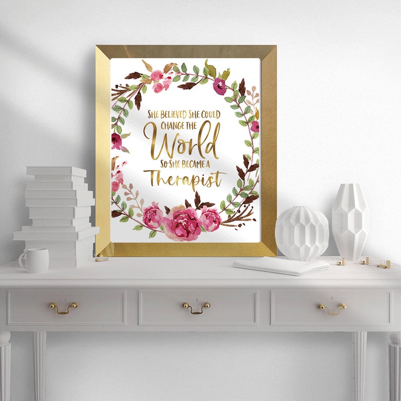 May include: A framed print with gold trim features the text "She believed she could change the world so she became a Therapist." The text is surrounded by a watercolor floral wreath in pink, burgundy, and green. The print is displayed on a white table.