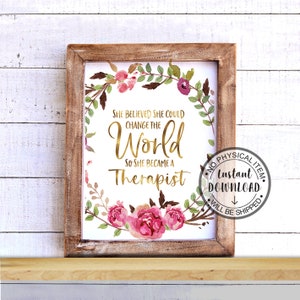 May include: A framed print with the text "She believed she could change the world so she became a therapist." The text is in gold script, surrounded by a floral wreath with pink and purple flowers and green leaves. The frame is made of light brown wood.