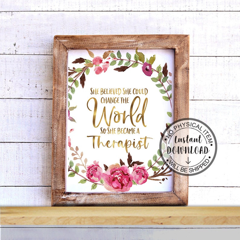 May include: A framed print with the text "She believed she could change the world so she became a therapist." The text is in gold script, surrounded by a floral wreath with pink and purple flowers and green leaves. The frame is made of light brown wood.