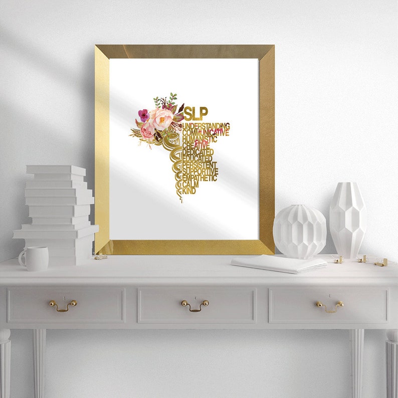May include: A gold framed print with a white background and a gold caduceus symbol with the word "SLP" at the top and a list of words describing the profession, such as "understanding", "communicative", "humanistic", "creative", "dedicated", "educated", "persistent", "supportive", "empathetic", "calm", and "kind". The caduceus is adorned with pink and white watercolor flowers.