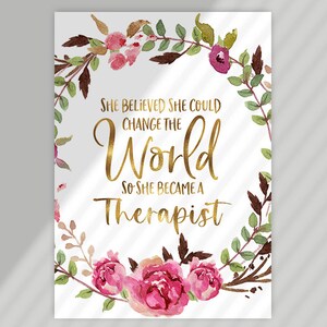 May include: A white print with the text "SHE BELIEVED SHE COULD CHANGE THE WORLD SO SHE BECAME A THERAPIST" in gold lettering, surrounded by a watercolor floral wreath in pink, green, and brown.