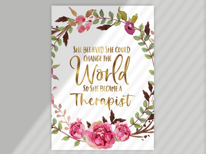 May include: A white print with the text "SHE BELIEVED SHE COULD CHANGE THE WORLD SO SHE BECAME A THERAPIST" in gold lettering, surrounded by a watercolor floral wreath in pink, green, and brown.