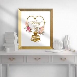 Op de afbeelding: Gouden ingelijst print met een gouden hart en de tekst "teaching is a work of heart". Het hart is omringd door roze en witte bloemen en een gouden appel op een stapel boeken.