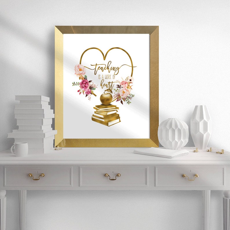 Op de afbeelding: Gouden ingelijst print met een gouden hart en de tekst "teaching is a work of heart". Het hart is omringd door roze en witte bloemen en een gouden appel op een stapel boeken.