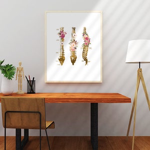 Puede incluir: Una impresi&oacute;n enmarcada de una ilustraci&oacute;n anat&oacute;mica dorada de la columna vertebral humana con acentos florales. La columna vertebral est&aacute; dividida en tres secciones, cada una con un arreglo floral diferente. La impresi&oacute;n est&aacute; sobre un fondo blanco y tiene un marco de madera simple.