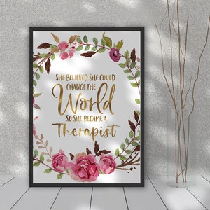 May include: A framed print with the text "She believed she could change the world so she became a therapist." The text is in gold against a white background, surrounded by a floral wreath with pink and burgundy flowers and green leaves.