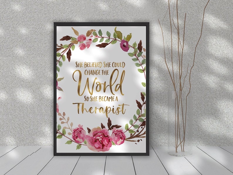 May include: A framed print with the text "She believed she could change the world so she became a therapist." The text is in gold against a white background, surrounded by a floral wreath with pink and burgundy flowers and green leaves.