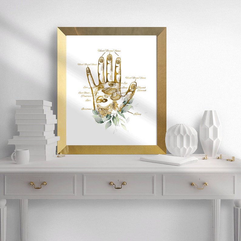 May include: A framed print featuring a golden hand with labeled palmistry details. The artwork includes botanical accents and is set in a gold frame. The print is displayed on a white surface with decorative items.