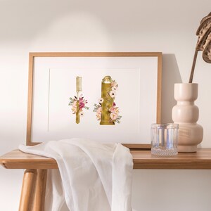 May include: A framed print of a gold toothbrush and toothpaste tube with floral accents. The print is on a white background and is framed in a natural wood frame. The print is sitting on a wooden table with a white cloth and a glass of water.