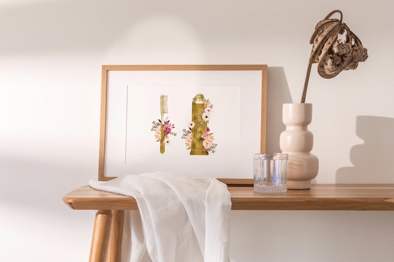 May include: A framed print of a gold toothbrush and toothpaste tube with floral accents. The print is on a white background and is framed in a natural wood frame. The print is sitting on a wooden table with a white cloth and a glass of water.