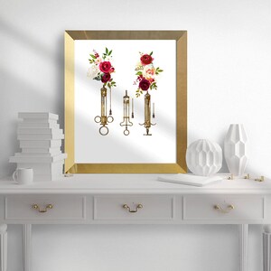 May include: A gold framed print with a white background featuring three vintage syringes with red and white floral arrangements at the top of each syringe.