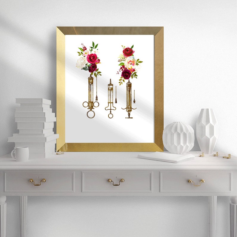 May include: A gold framed print with a white background featuring three vintage syringes with red and white floral arrangements at the top of each syringe.