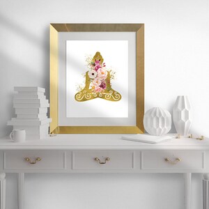Op de afbeelding: Een gouden silhouet van een mediterende figuur met roze en witte bloemen in de figuur. De afbeelding is ingelijst in een gouden lijst en staat op een witte plank met witte decoratie.