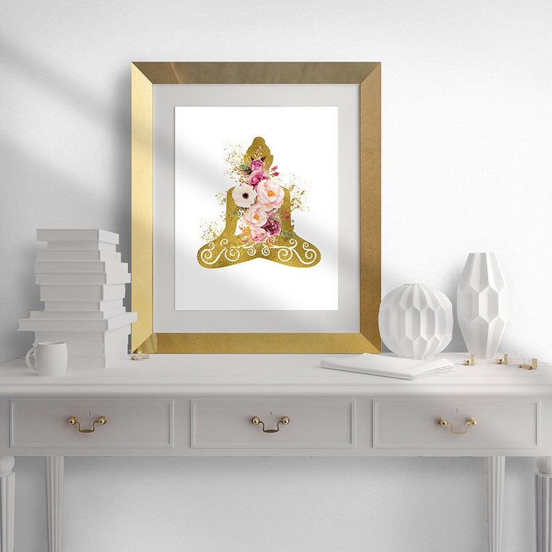 Op de afbeelding: Een gouden silhouet van een mediterende figuur met roze en witte bloemen in de figuur. De afbeelding is ingelijst in een gouden lijst en staat op een witte plank met witte decoratie.