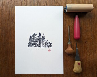 May include: A black and white line drawing of Notre Dame de Paris, a famous cathedral in Paris, France. The drawing is on a white sheet of paper with a red stamp in the bottom right corner. The image also shows tools used for printmaking, including a wooden roller, a carving tool, and a small wooden handle with a metal blade.