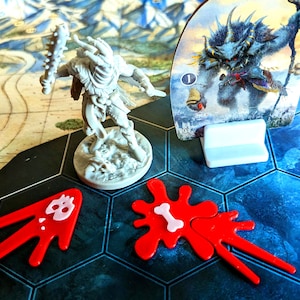 May include: A board game scene featuring a miniature warrior figure with a spiked club, a monster card, and red game pieces resembling blood splatters. The game board has a map and hexagonal tiles.