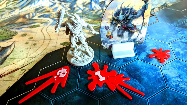 May include: A board game scene featuring a miniature warrior figure with a spiked club, a monster card, and red game pieces resembling blood splatters. The game board has a map and hexagonal tiles.