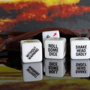 May include: A set of white dice with black text, including phrases like "ROLL SOME DICE" and "SHAKE HEAD SADLY." The dice are displayed with a brown drawstring bag against a blurred, colorful background.