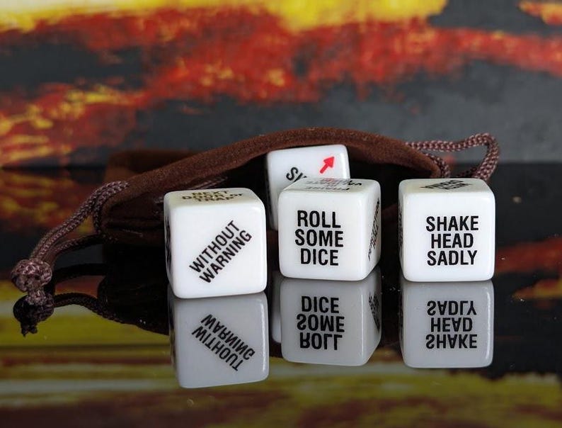 May include: A set of white dice with black text, including phrases like "ROLL SOME DICE" and "SHAKE HEAD SADLY." The dice are displayed with a brown drawstring bag against a blurred, colorful background.
