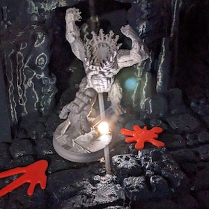 May include: A gray figurine of a muscular figure with a crown, posed in a dramatic stance. The figure is on a round base, illuminated by a small light. Two red, abstract shapes are on the dark, textured surface.